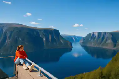 A visitor sitting on the rail of the viewpoint above a long blue fjord
